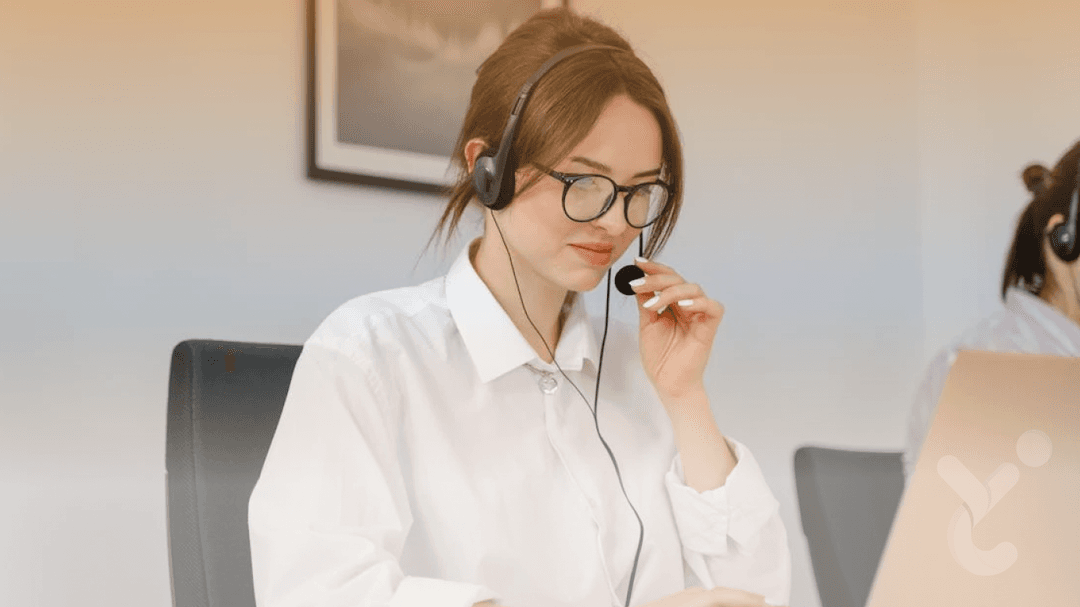 A woman wearing glasses and a white shirt, using a headset microphone while working on a laptop, sitting in an office environment.