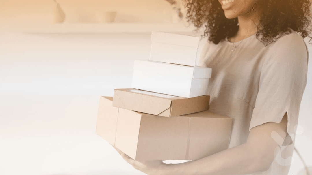 A smiling woman holding a stack of cardboard and white boxes, standing indoors in a minimalistic setting, representing order management or delivery services.